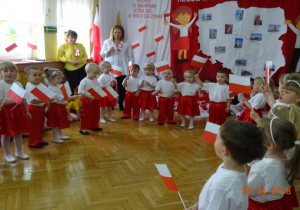 Na tle dekoracji z mapą Polski i dziećmi z chorągiewkami stoją w półkolu z z chorągiewkami.